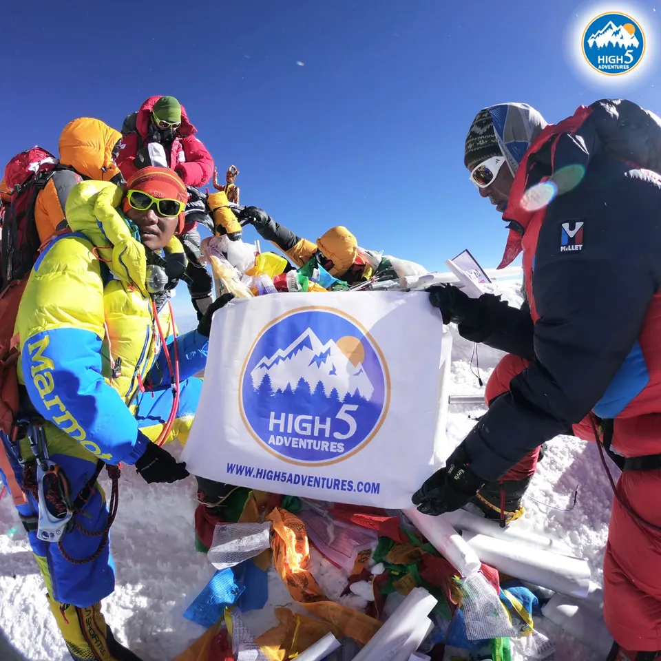 Two climbers holding a High 5 Adventures banner among prayer flags, on the summit of Everest