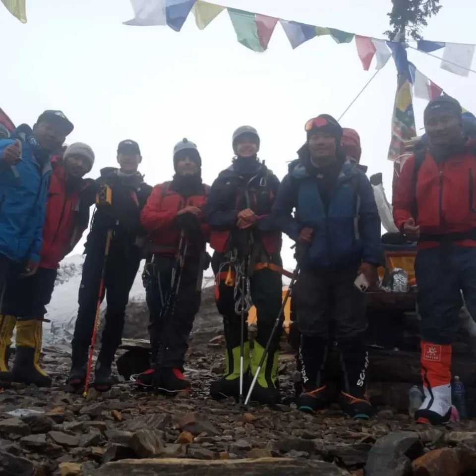 Seven climbers in high-altitude boots and harnesses under prayer flags, Manaslu Base Camp