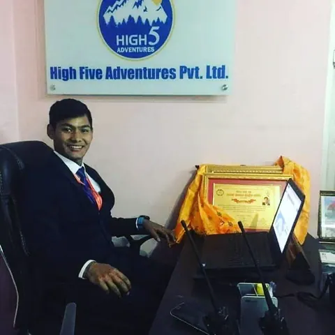 A man in a dark suit at a desk with framed certificates, below the High Five Adventures Pvt. Ltd. sign in the company office
