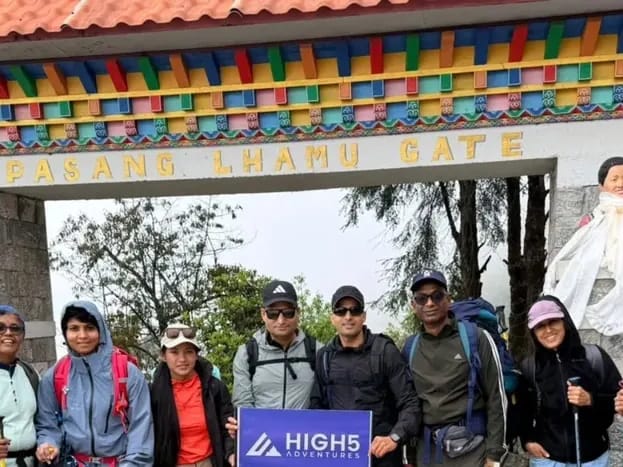 Daypacks and trekking poles at the start in Lukla: The trek group at the Pasang Lhamu Gate in Lukla, May 2026. Trekkers holding a High 5 Adventures banner under the Pasang Lhamu Gate in Lukla, May 2026. Trekkers holding a High 5 Adventures banner under the Pasang Lhamu Gate in Lukla, May 2026
