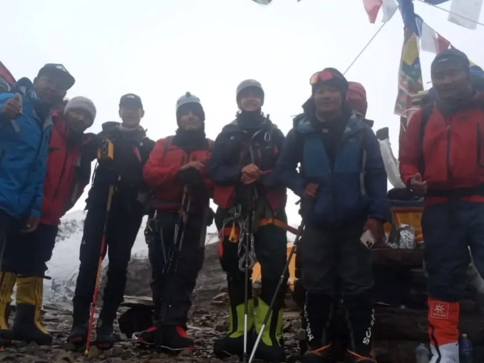 Seven climbers in high-altitude boots and harnesses under prayer flags, Manaslu Base Camp