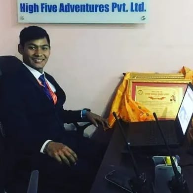 A man in a dark suit at a desk with framed certificates, below the High Five Adventures Pvt. Ltd. sign in the company office