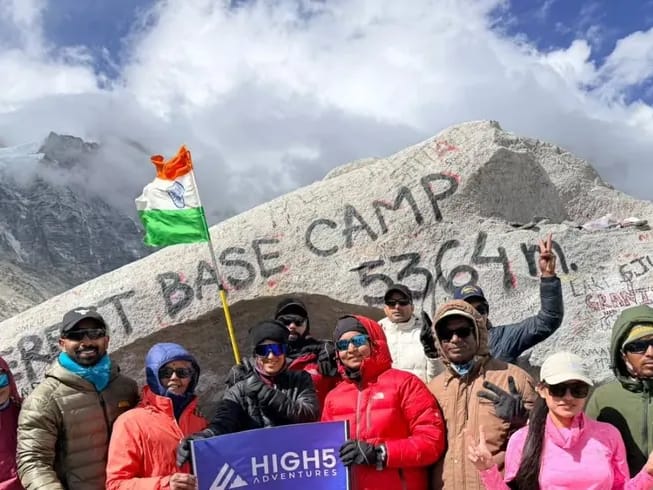 The Everest Base Camp trek: The May 2026 Everest Base Camp group with the High 5 banner. Trekkers holding a High 5 Adventures banner, with an Indian flag, in front of the painted 5,364 m boulder at Everest Base Camp, May 2026. Trekkers holding a High 5 Adventures banner, with an Indian flag, in front of the painted 5,364 m boulder at Everest Base Camp, May 2026