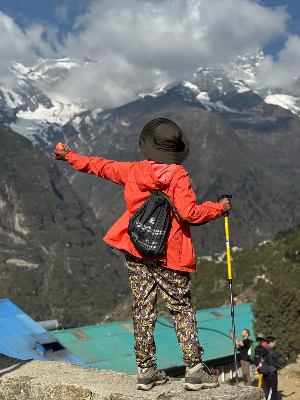 A trekker in an orange jacket and wide-brimmed hat on a stone wall, arms spread wide, above the tin roofs of Namche Bazaar, with snow peaks in cloud beyond, May 2026