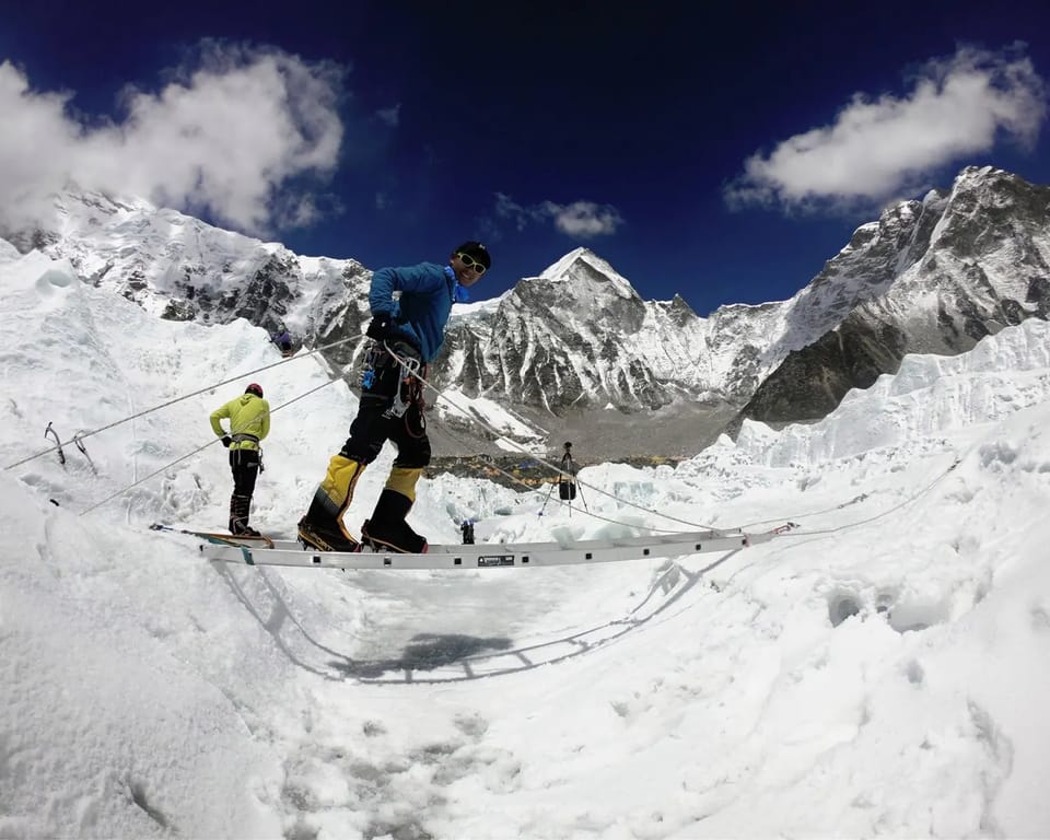 A climber in a blue jacket standing on an aluminium ladder across a crevasse, clipped to fixed ropes, with a tented camp below